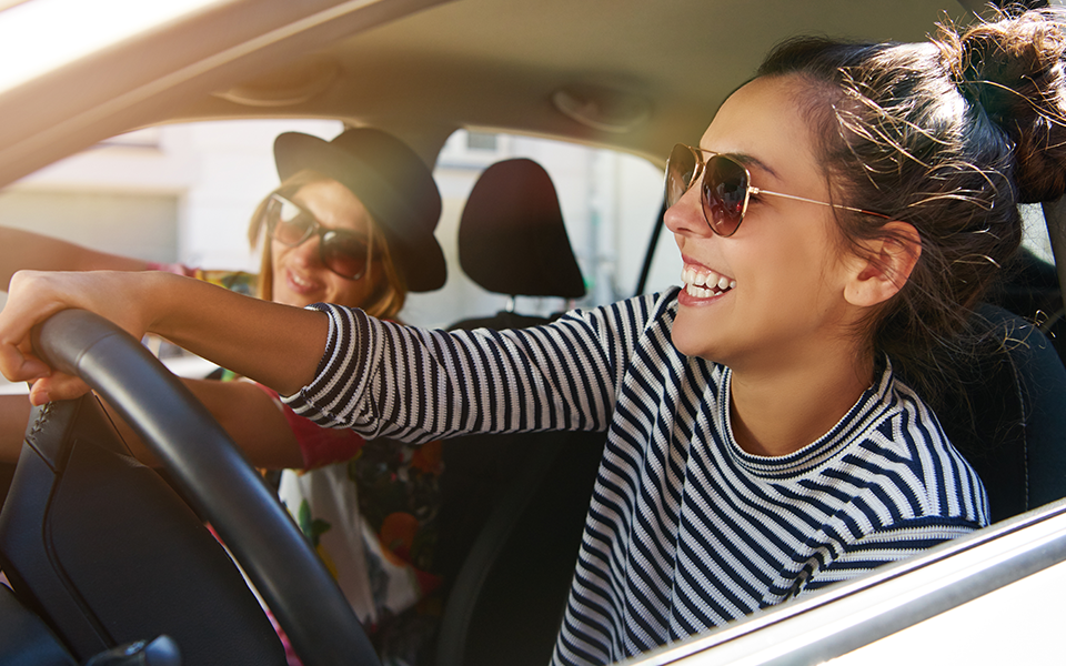 A woman is smiling in the driver's seat of a car. She appears to be driving and she has a friend seated in the passenger seat with her