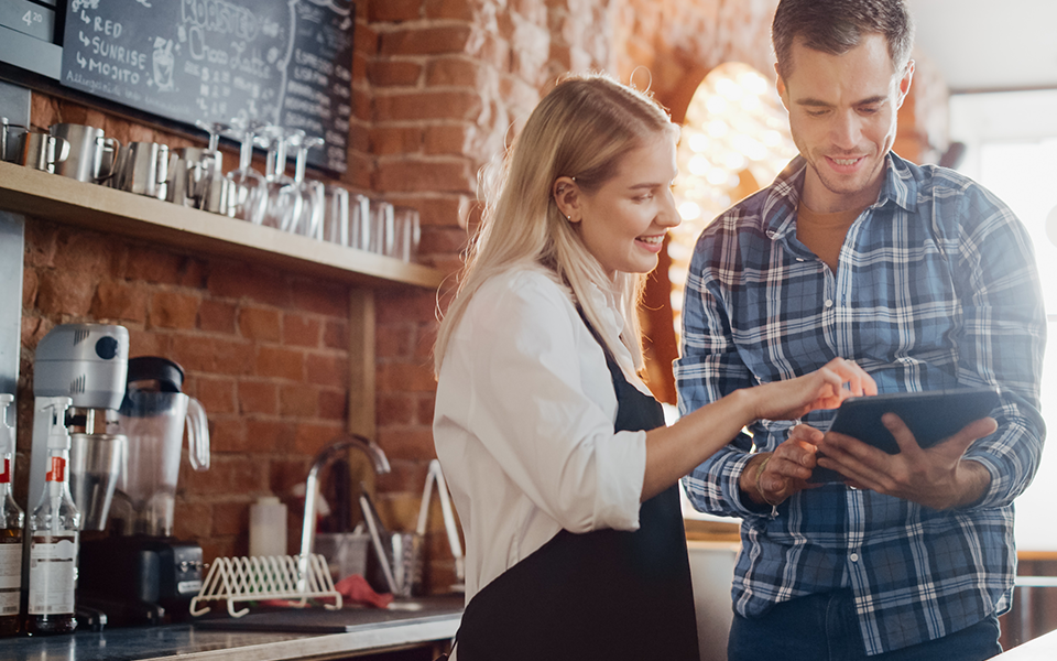 A man in a plaid flannel shirt holds a tablet. Next to him, a woman in an apron is pointing to something on the tablet's screen