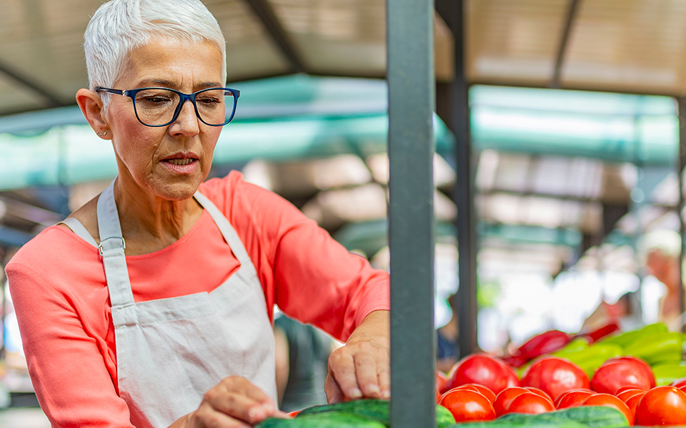 An woman with close-cropped gray hair is working at a produce stand wearing a light colored apron over her coral shirt. She is looking at produce