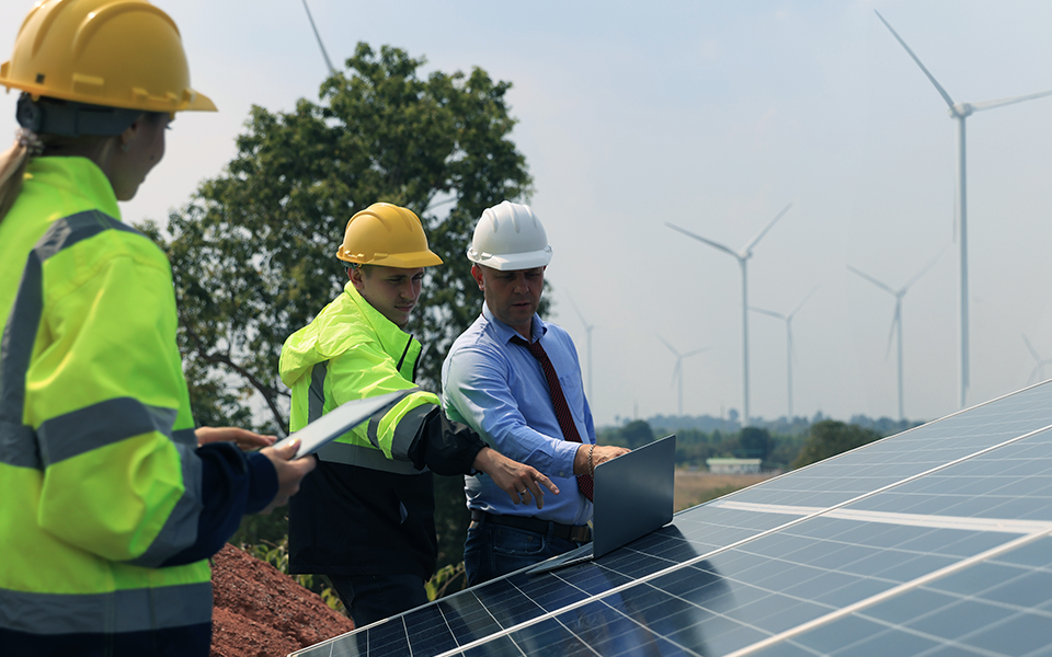Business-Commercial-Loan-960x600 – Capital Bank Two workers and one business man are looking at a laptop while working on solar panels. All three are wearing hard hats.