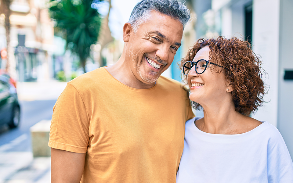 A husband and wife are outside walking on a sidewalk. She is looking at him and he's smiling