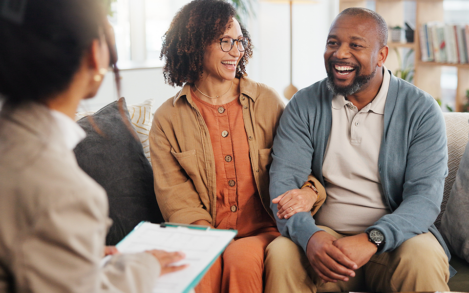 Financial-Planning-960x600 – Capital Bank A couple seated closely on a sofa are laughing. They appear to be working with an advisor who is facing them and is taking notes on a clipboard