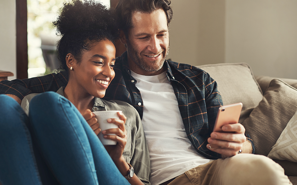 A man and woman sit closely on the couch. He has his arm around her. She is holding a cup of coffee and he is holding his phone and they are both looking at the screen