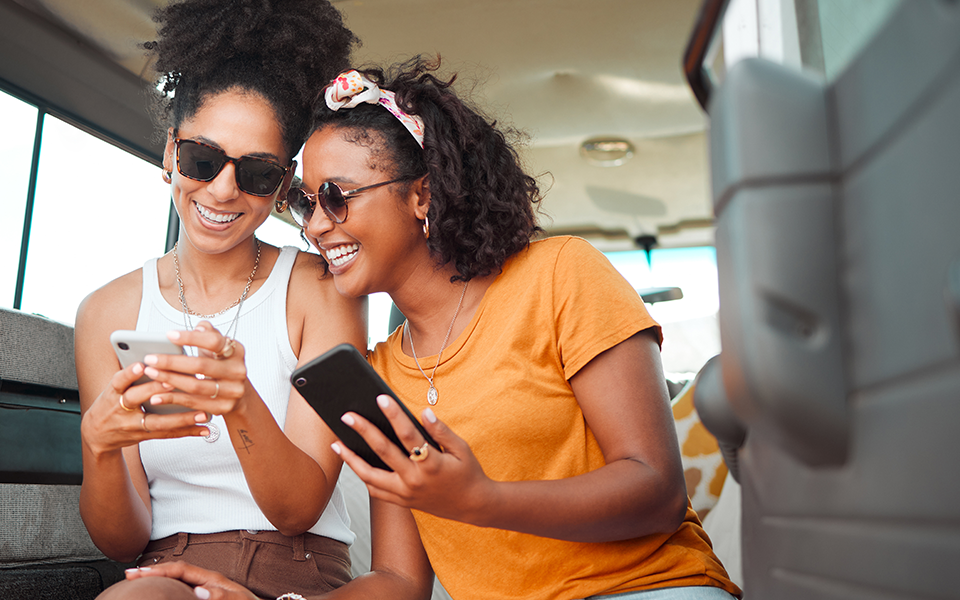 Personal-Checking-960x600 – Capital Bank Two friends are seated near each other. Both women are holding smart phones. The woman on the right is leaning over and smiling at the phone screen of the woan on the left