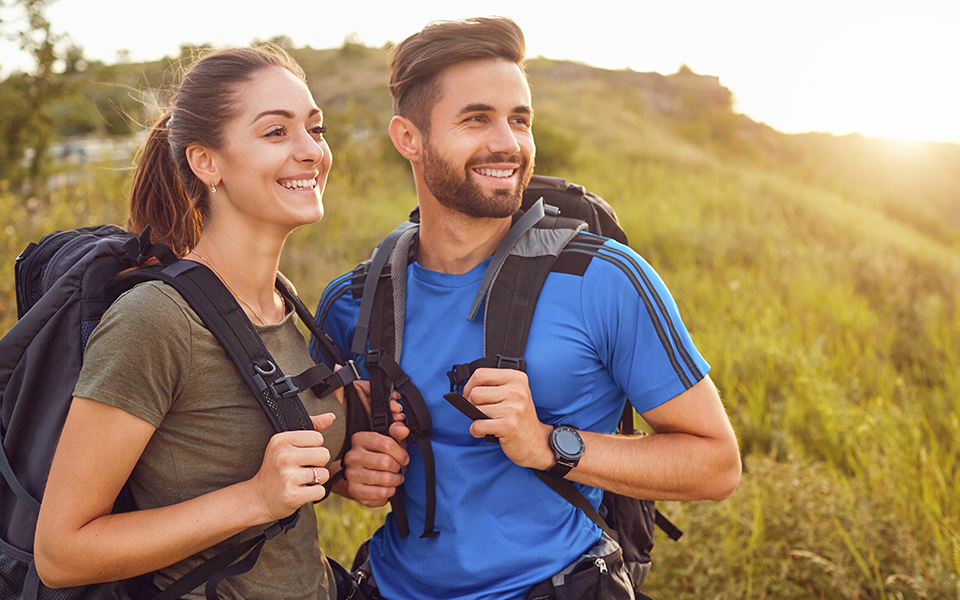 A man and woman are outside in the late golden sun of an afternoon. They are both wearing hiking backpacks and looking happily off in the distance
