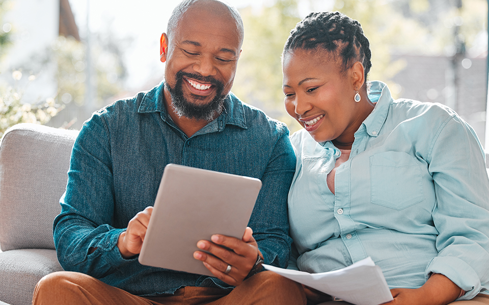 A husband and wife are seated closely on a sofa. He is holding a tablet in his hand and they appear to be talking about what he's inputting on the screen