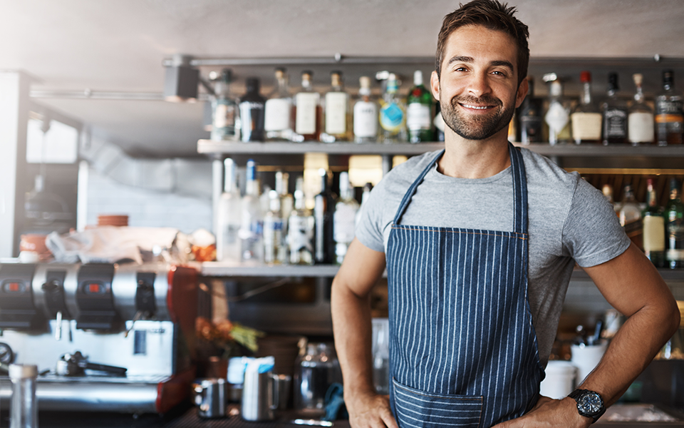 A man is standing in a cafe in front of shelves of bottled liquors and syrups. An espresso machine is behind him