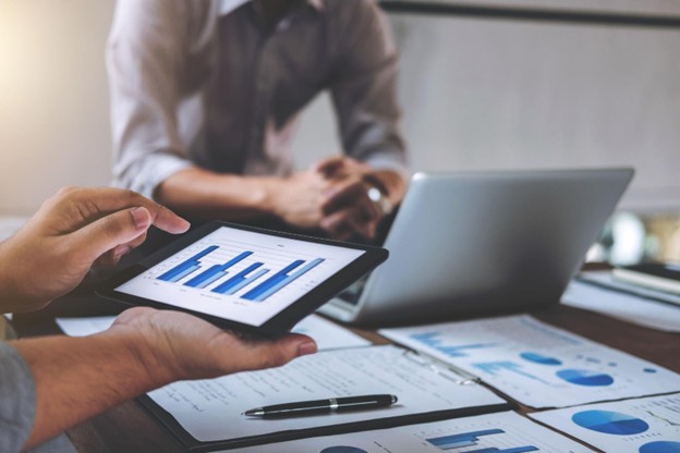 Close up of an image of two people working at a table with a tablet in front of one, and a laptop in front of the other, and various documents on the table in in front of them