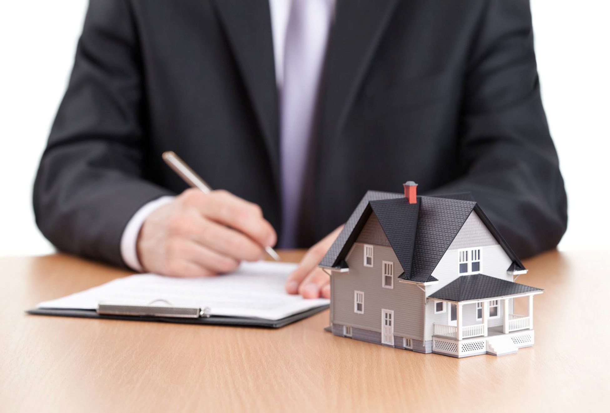 Photo of a man in a business suit writing on a notepad. There is a model of a house on the table in front of his notebook.