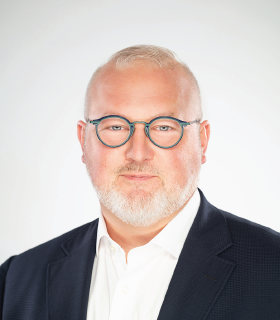 Portrait of a middle-aged man with gray hair and beard wearing rectangular glasses, a white shirt, and a dark blazer, looking at the camera against a light background.