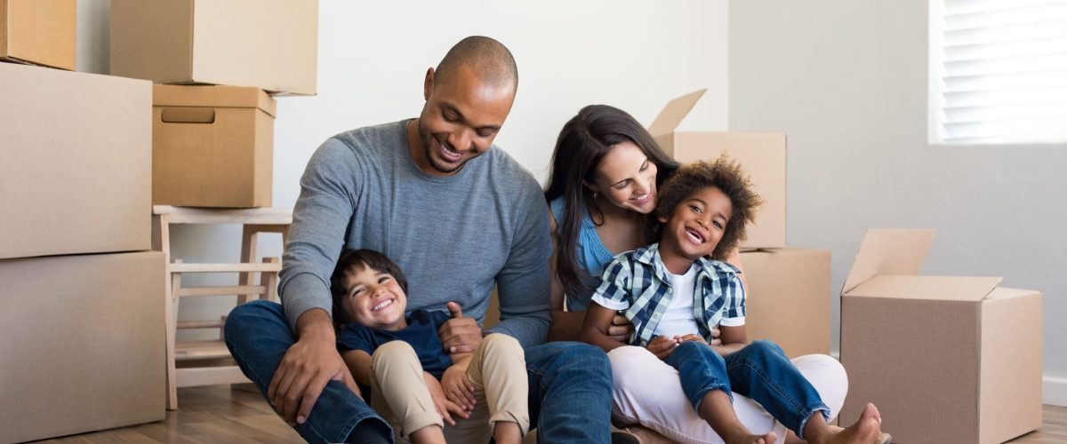 Happy family with two children having fun at new home. Young multiethnic parents with two sons in their new house with cardboard boxes. Smiling little boys sitting on floor with mother and dad.