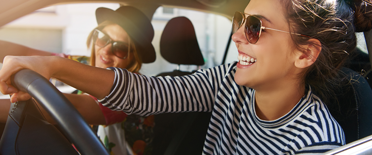 A woman is smiling in the driver's seat of a car. She appears to be driving and she has a friend seated in the passenger seat with her