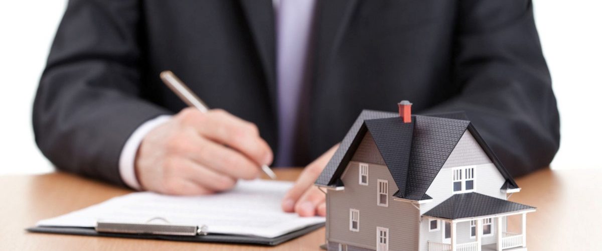 Photo of a man in a business suit writing on a notepad. There is a model of a house on the table in front of his notebook.