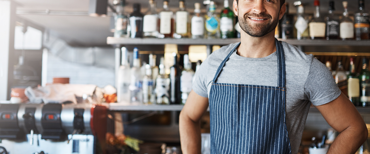A man is standing in a cafe in front of shelves of bottled liquors and syrups. An espresso machine is behind him
